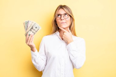 Güzel sarışın kız düşünüyor, kuşkulu ve şaşkın hissediyor. Dolar banknotları konsepti