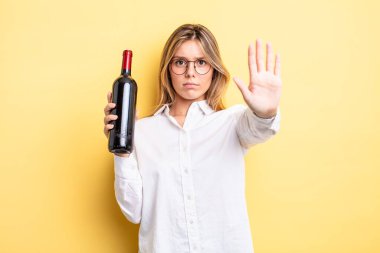 Güzel sarışın kız ciddi görünüyor. Avuç içini gösteriyor. Durma hareketi yapıyor. şarap şişesi konsepti