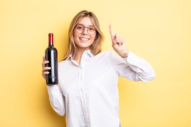 Güzel sarışın kız gülümsüyor ve arkadaş canlısı görünüyor. Bir numarayı gösteriyor. şarap şişesi konsepti
