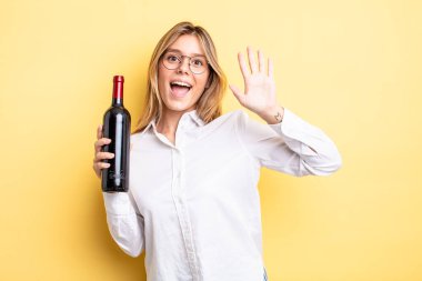 Güzel sarışın kız mutlu bir şekilde gülümsüyor, el sallıyor, hoş geldin diyor ve seni selamlıyor. şarap şişesi konsepti
