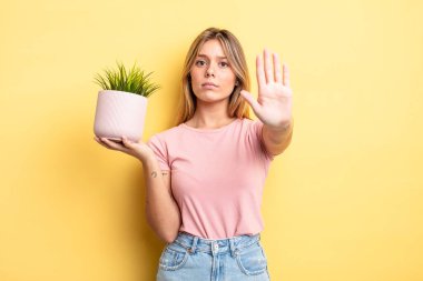 Güzel sarışın kız ciddi görünüyor. Avuç içini gösteriyor. Durma hareketi yapıyor. ev bitkisi kavramı