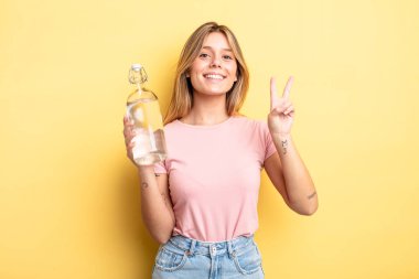 Güzel sarışın kız gülümsüyor ve arkadaş canlısı görünüyor. İki numarayı gösteriyor. su şişesi kavramı
