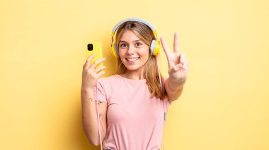 Güzel sarışın kız gülümsüyor ve mutlu görünüyor, zafer ya da barış işareti yapıyor. müzik konseptini dinlemek