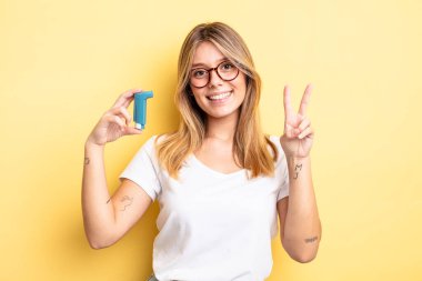 Güzel sarışın kız gülümsüyor ve arkadaş canlısı görünüyor. İki numarayı gösteriyor. astım spreyi kavramı