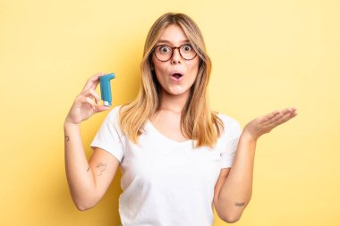 Güzel sarışın kız şaşırmış ve şok olmuş görünüyor. Çenesi düşmüş ve elinde bir nesne var. astım spreyi kavramı