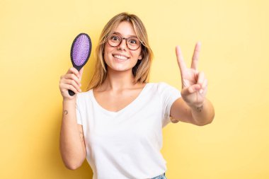 Güzel sarışın kız gülümsüyor ve mutlu görünüyor, zafer ya da barış işareti yapıyor. saç fırçası konsepti