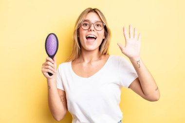 Güzel sarışın kız mutlu bir şekilde gülümsüyor, el sallıyor, hoş geldin diyor ve seni selamlıyor. saç fırçası konsepti