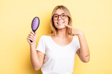 Güzel sarışın kız kendi gülüşünü göstererek kendine güvenerek gülümsüyor. saç fırçası konsepti