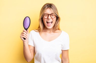 Güzel sarışın kız agresif bir şekilde bağırıyor, çok kızgın görünüyor. saç fırçası konsepti