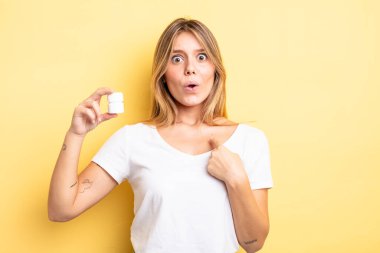 Şok olmuş ve ağzı açık, kendini işaret eden güzel sarışın bir kız. hap şişesi konsepti