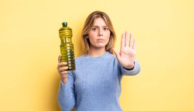 Güzel sarışın kız ciddi görünüyor. Avuç içini gösteriyor. Durma hareketi yapıyor. Zeytin yağı kavramı