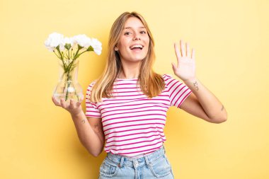 Güzel sarışın kız mutlu bir şekilde gülümsüyor, el sallıyor, hoş geldin diyor ve seni selamlıyor. Dekoratif çiçek konsepti