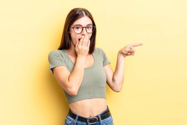 İspanyol güzel kadın kendini mutlu, şaşkın ve şaşırmış hissediyor. Ağzını eliyle kapatıyor ve yanal kopya alanını işaret ediyor.