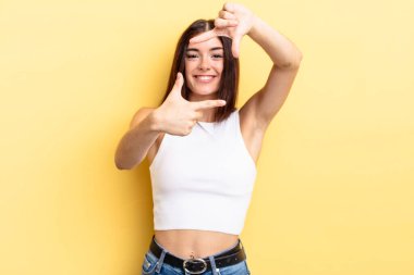 İspanyol güzel kadın mutlu, arkadaş canlısı ve pozitif hissediyor, gülümsüyor ve elleriyle bir portre veya fotoğraf çerçevesi yapıyor.