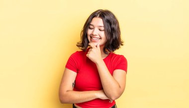 Genç ve güzel bir kız, mutlu, kendinden emin bir ifadeyle gülümsüyor, elleri çenesinde, merak ediyor ve yan tarafa bakıyor.