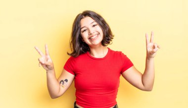 Genç ve güzel bir kız gülümsüyor ve mutlu görünüyor, arkadaş canlısı ve memnun, işaret eden zafer ya da huzur iki elinle