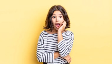 Genç ve güzel bir kız şok ve inançsızlık içinde, elleri yanağında ve kolları çaprazlanmış, sersemlemiş ve şaşırmış hissediyor.