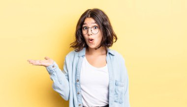 Genç ve güzel bir kız şaşırmış ve şok olmuş görünüyor, çenesi düşmüş, bir objeyi elinde açık bir elle tutuyor.
