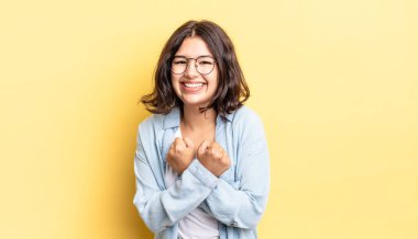 Genç ve güzel bir kız, neşeli bir şekilde gülümsüyor ve kutluyor. Yumruklarını sıkmış, kollarını kavuşturmuş, mutlu ve pozitif hissediyor.