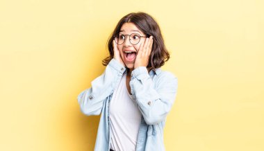 Genç, güzel bir kız mutlu, heyecanlı ve şaşırmış hissediyor, iki eli yüzü üzerinde yana bakıyor.