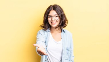 Genç, güzel bir kız gülümsüyor, mutlu, kendinden emin ve arkadaş canlısı görünüyor, bir anlaşmaya varmak için el sıkışmayı teklif ediyor, işbirliği yapıyor.