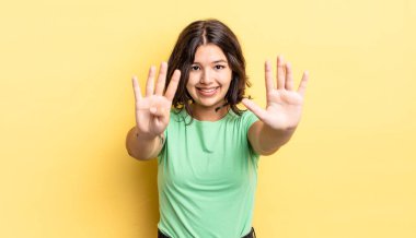 Genç, güzel bir kız gülümsüyor ve arkadaş canlısı görünüyor. 9 ya da 9 numarayı gösteriyor. Elleri önde ve geri sayıyor.