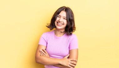 Genç ve güzel bir kız kollarını kavuşturup mutlu bir şekilde gülüyor, rahat, pozitif ve tatmin edici bir pozla.