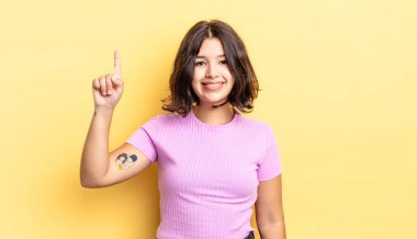 Genç ve güzel bir kız mutlu bir şekilde gülümsüyor, tek eliyle yukarıyı işaret ederek boşluğu kopyalıyor.