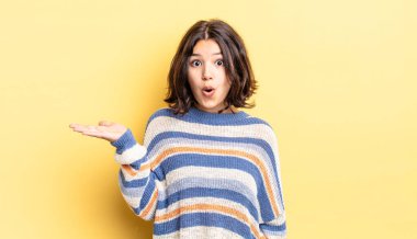 Genç ve güzel bir kız şaşırmış ve şok olmuş görünüyor, çenesi düşmüş, bir objeyi elinde açık bir elle tutuyor.