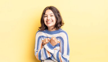 Genç ve güzel bir kız, neşeli bir şekilde gülümsüyor ve kutluyor. Yumruklarını sıkmış, kollarını kavuşturmuş, mutlu ve pozitif hissediyor.