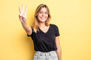 Sarışın, güzel bir kadın gülümsüyor ve arkadaş canlısı görünüyor. Üç ya da üç numarayı gösteriyor. Elleri önde, geriye doğru sayıyor.