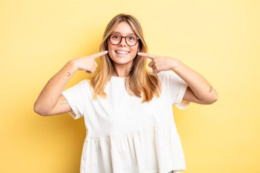 Sarışın, güzel bir kadın, kendine güvenerek gülümsüyor. Geniş bir gülümseme, pozitif, rahat, tatmin olmuş bir tavır.