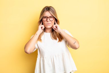 Sarışın, güzel bir kadın sinirli, stresli ve sinirli görünüyor, iki kulağını da sağır edici bir ses, ses ya da yüksek sesle kapatıyor.