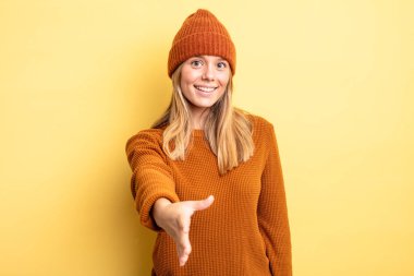 Sarışın, güzel bir kadın gülümsüyor, mutlu, kendinden emin ve arkadaş canlısı görünüyor, bir anlaşmaya varmak için el sıkışmayı teklif ediyor, işbirliği yapıyor.