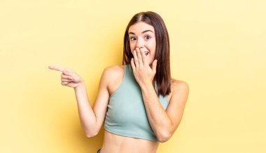 İspanyol güzel kadın kendini mutlu, şaşkın ve şaşırmış hissediyor. Ağzını eliyle kapatıyor ve yanal kopya alanını işaret ediyor.