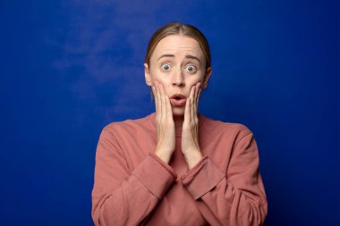 Sarışın kadın şok olmuş ve korkmuş hissediyor, ağzı açık ve elleri yanaklarında korkmuş görünüyor.