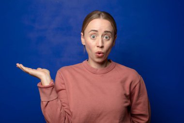 Sarışın kadın şaşırmış ve şoka uğramış görünüyor. Çenesi düşmüş ve eli açık bir objeyi tutuyor.