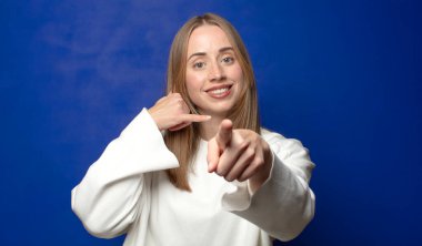 Neşeli bir şekilde gülümsüyor ve telefonla konuşurken kamerayı işaret ediyor.