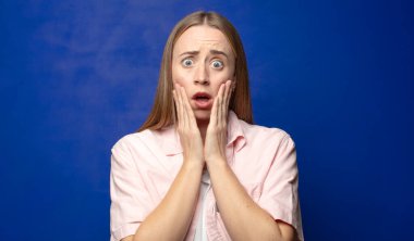 Şok olmuş ve korkmuş hissediyorum, ağzı açık ve elleri yanaklarında korkmuş görünüyor.