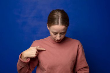 Sarışın kadın neşeyle gülümsüyor, yere bakıyor ve göğsünü işaret ediyor.