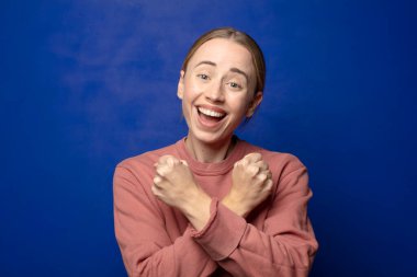 Sarışın kadın sevinçle gülümsüyor ve kutluyor, yumruklarını sıkmış ve kollarını kavuşturmuş, mutlu ve pozitif hissediyor.