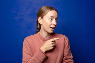 Şok olmuş ve şaşırmış hisseden sarışın bir kadın, şaşkınlık içinde, ağzı açık bir bakışla yukarı bakıyor ve onu işaret ediyor.