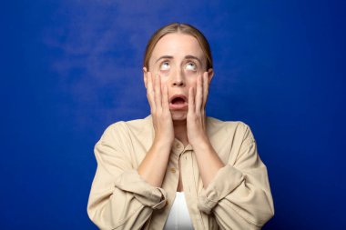 Sarışın kadın şok olmuş ve korkmuş hissediyor. Elleri yanaklarında sıkılmış görünüyor.