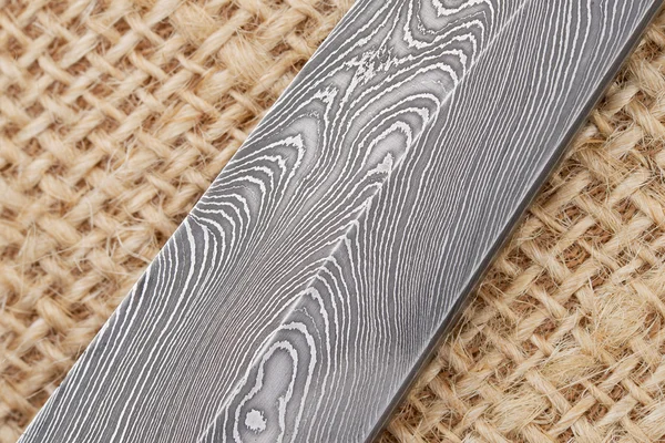 Fragment of the traditional handmade Finnish knife blade with the abstract wave pattern of damascus steel over an old sack background. — Stock Photo, Image