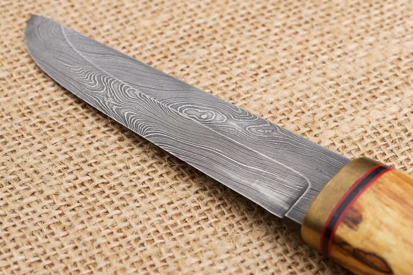 Traditional handmade Finnish knife with the abstract wave pattern of damascus steel and wooden handle over an old sack background. — Stock Photo, Image