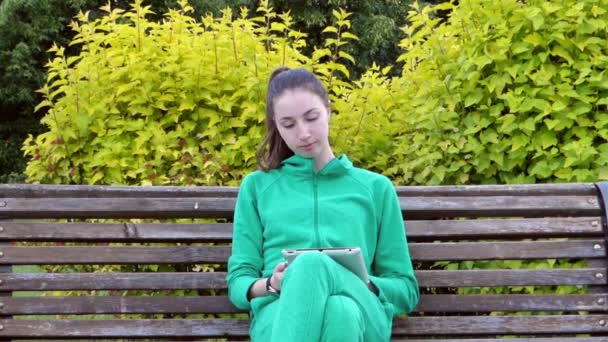 Joven y hermosa chica sentada en un banco en el parque y usar la tableta. Ella mira el reloj volteando a través de las noticias y se ríe . — Vídeos de Stock