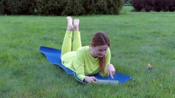 Menina desportiva depois de Yoga, deitado no estômago e usando seu tablet. Sorrindo. . — Vídeo de Stock