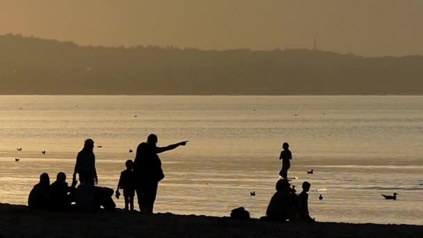 Gente descansando en la playa admirando la puesta de sol. Moción lenta . — Vídeo de stock