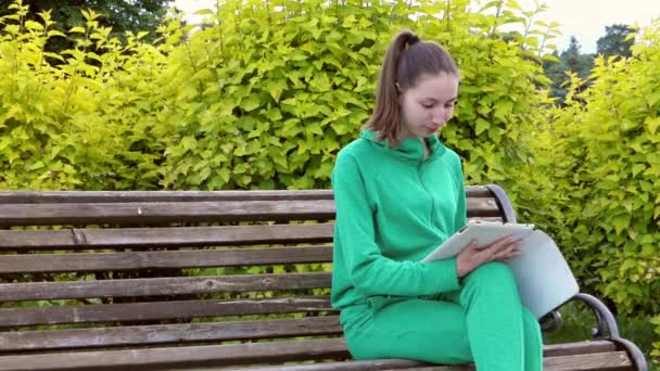 Mädchen sitzt auf einer Bank und scannt die Nachrichten auf dem Tablet. Sie genießt die neue Botschaft. — Stockvideo