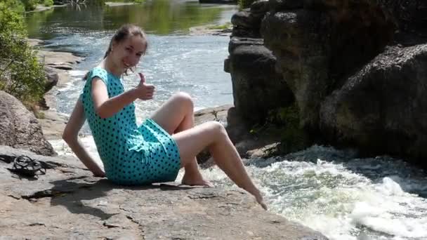 Menina bonita tem um descanso sentado em uma pedra perto de um rio da montanha. Ela molha seus pés e brinca com a água . — Vídeo de Stock